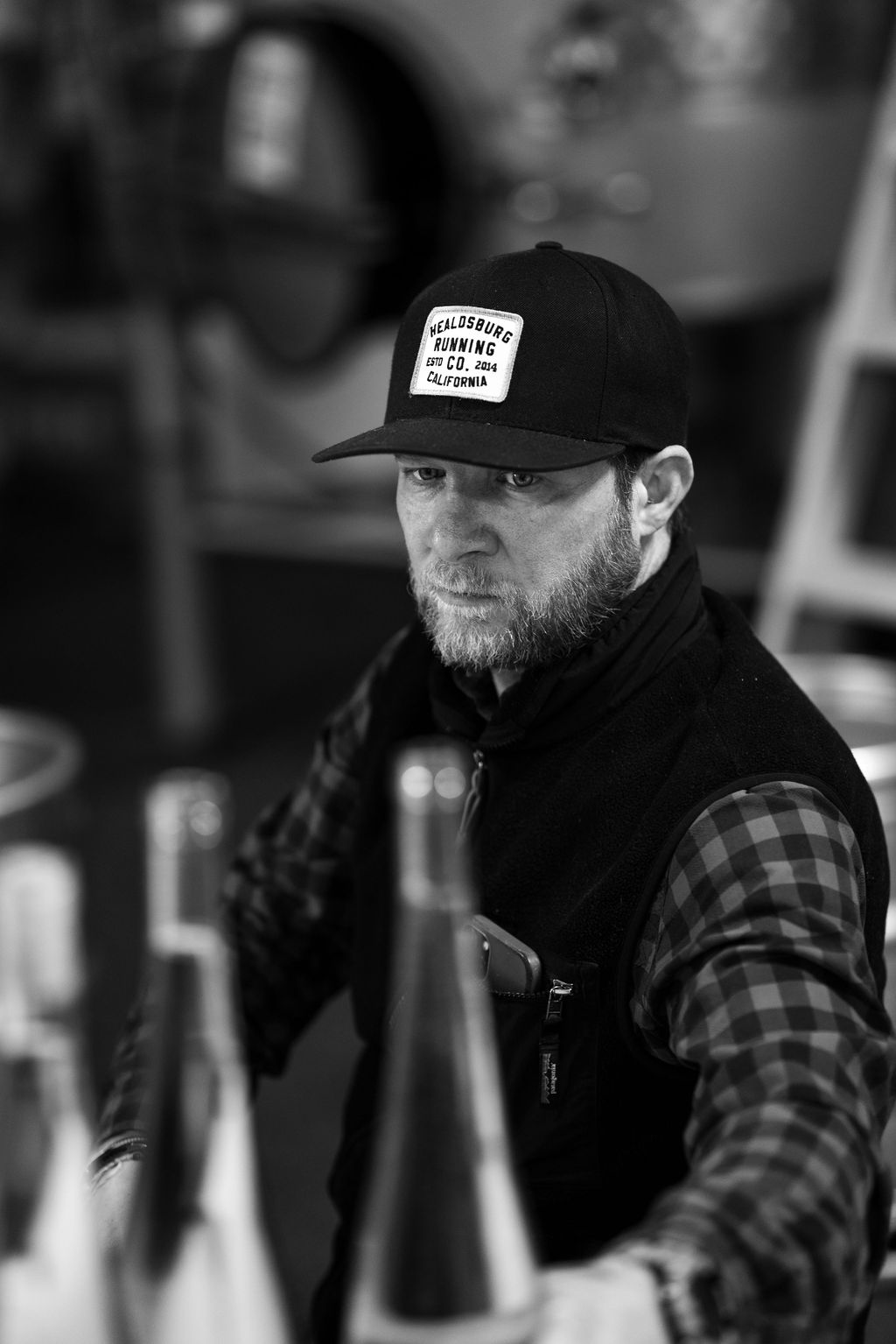 DSC01992 Black-and-white portrait of a bearded man in a cap among bottles.